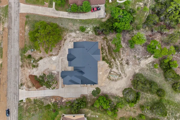 Top-down aerial of a charcoal metal roofline, curved concrete drive, and cleared homesite among oaks