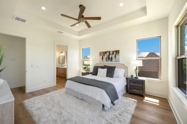 Primary suite with recessed tray ceiling, tall windows on two walls, and ensuite bath with wood vanity beyond