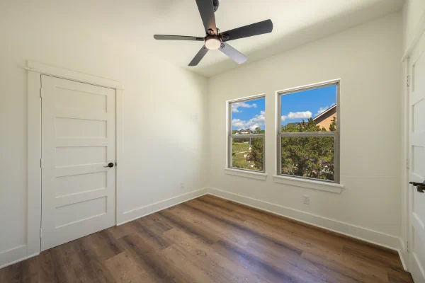 Empty bedroom with five-panel door, ceiling fan, and two windows overlooking cedar treetops