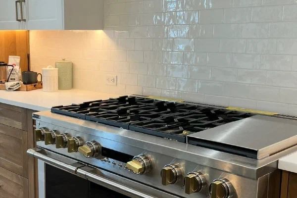 Professional gas range with brass knobs and griddle under a stained wood vent hood and glossy subway tile