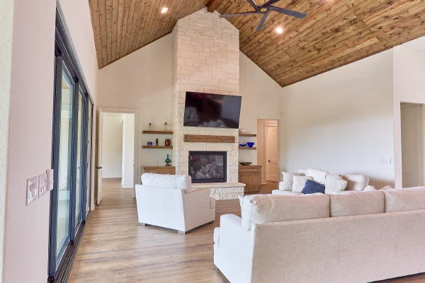Living room with wood plank cathedral ceiling, stone fireplace with mounted TV, and black-framed sliding glass doors