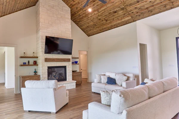 Neutral seating area facing a stone fireplace with cedar mantel beneath a stained pine vaulted ceiling