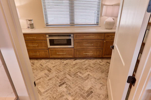 Butler's pantry with oak cabinetry, brass pulls, microwave drawer, and herringbone travertine tile floor
