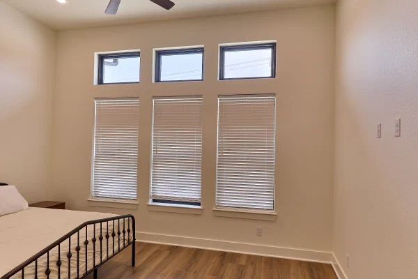 Bedroom with triple windows topped by transoms, faux wood blinds, iron bed frame, and wood-look plank flooring