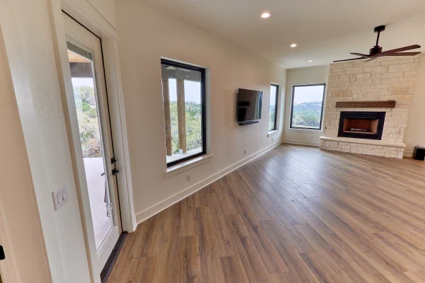 Game room with corner fireplace in cream limestone, rustic wood mantel, and picture windows framing wooded views