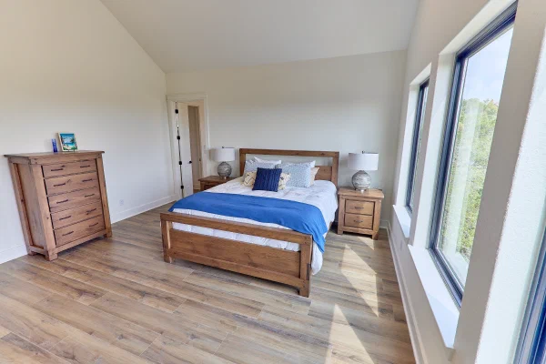 Primary bedroom with oak five-drawer chest, king bed with blue throw, and tall black-framed corner windows