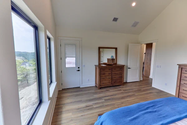 Primary suite with windows framing Texas Hill Country oaks, patio access door, and rustic oak dresser with mirror