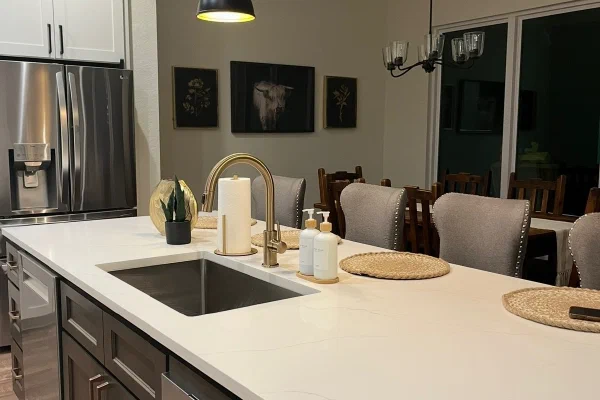 Evening-lit quartz island with gold faucet and dark wood cabinets beside an upholstered dining area