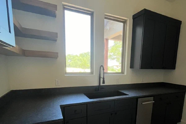 Matte black cabinetry with leathered stone counters, floating wood shelves and a herringbone brick floor