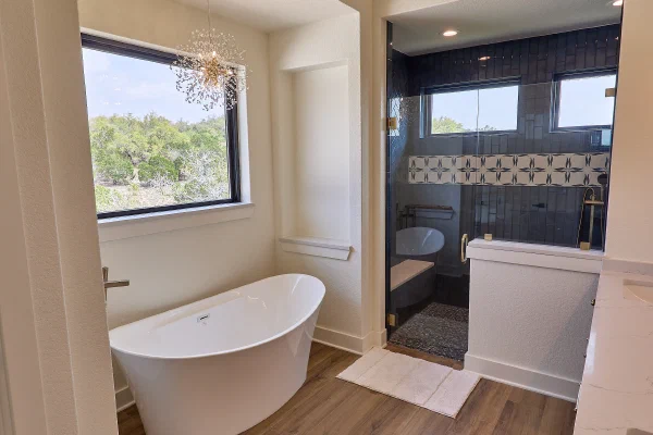 Freestanding oval tub beneath a window and crystal chandelier, with a glass-enclosed navy tile shower beyond