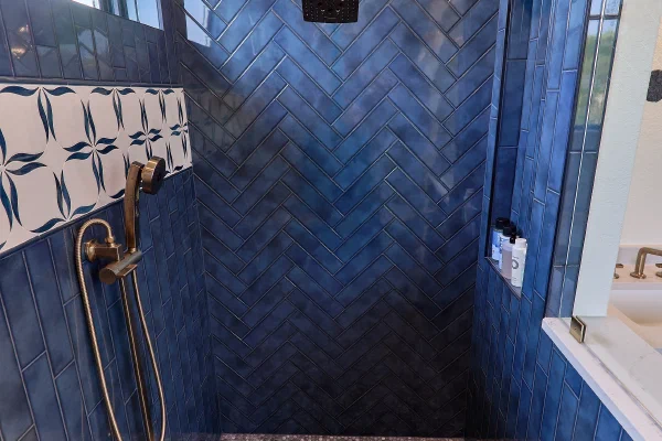 Walk-in shower with glossy blue herringbone tile, rain shower head, brass handheld sprayer, and tiled storage niche